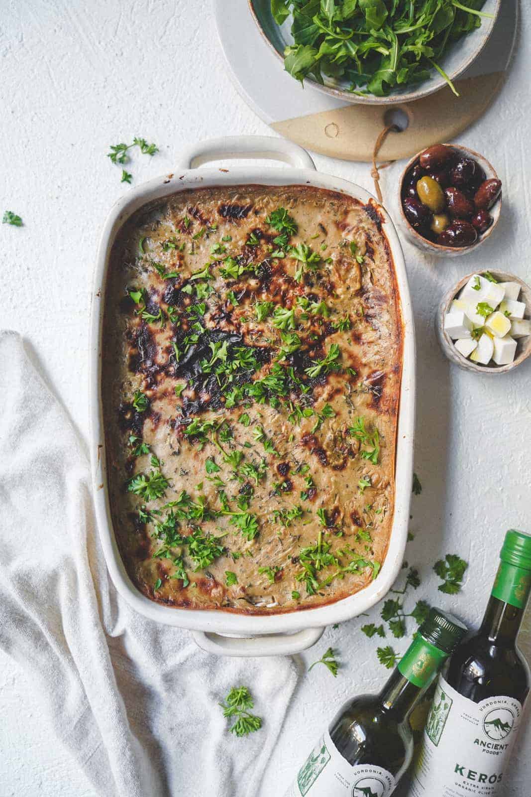 Vegan moussaka dish sitting on countertop ready to be cut into, surrounded by key Greek ingredients like vegan feta and olives.