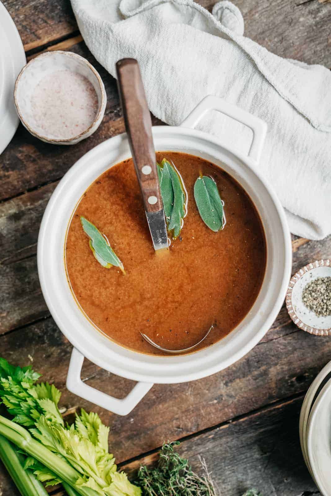 Bowl of homemade vegetable stock with fresh herbs on top on wooden countertop.