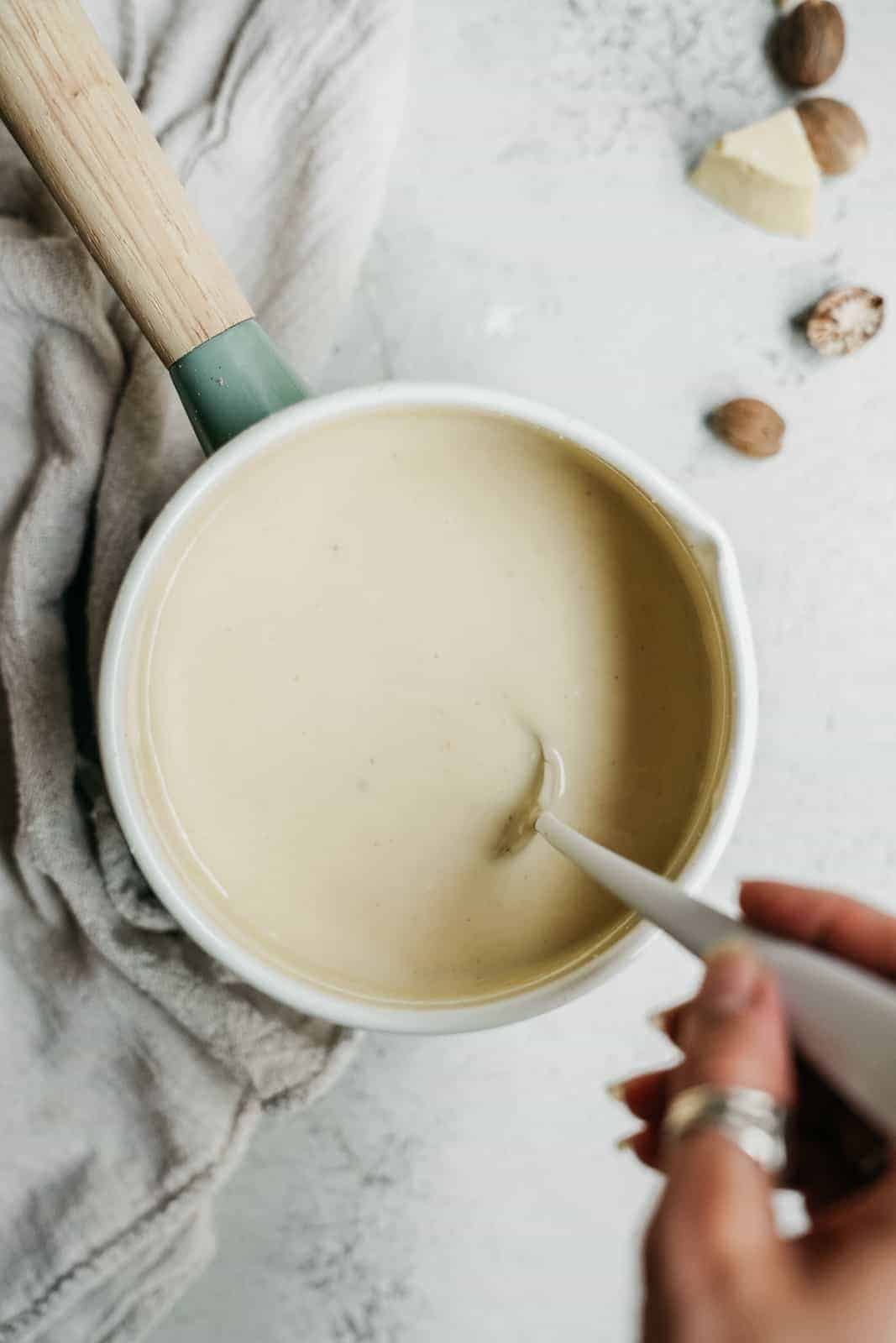 Pot with vegan bachamel sauce being stirred with a spoon.