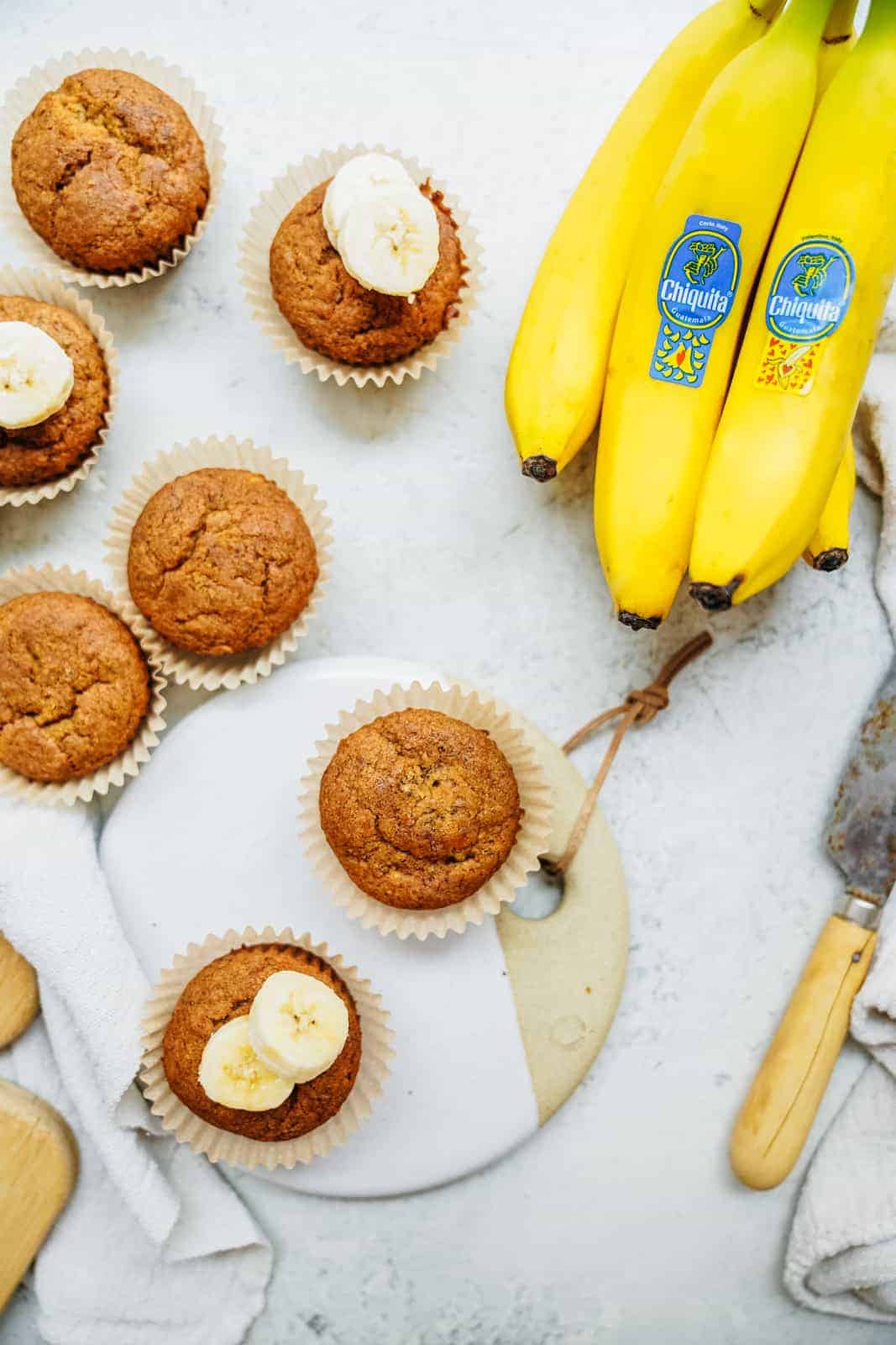 Quick and easy vegan banana bread muffins scattered on countertop with a bunch of fresh yellow bananas.