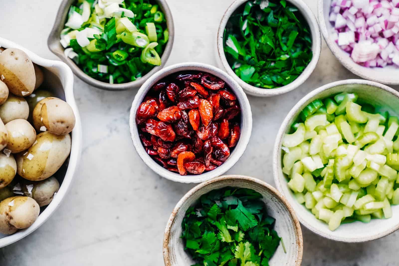 Fresh herbs and veggies with cooked potatoes and dried cranberries in bowls, ready to be mixed.