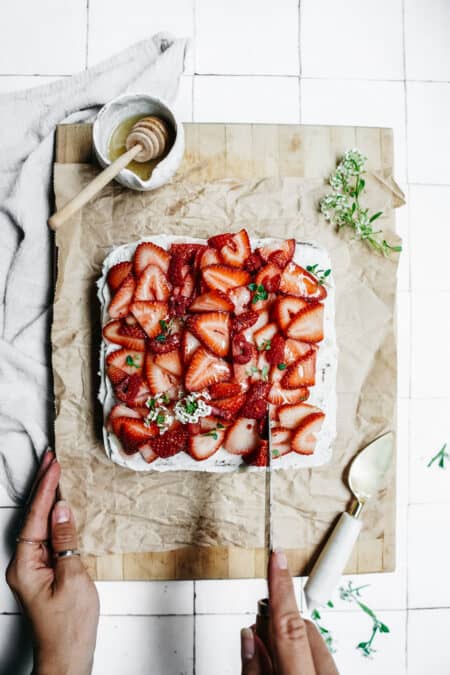 Slice of delicious Honey Cake made with BeeMaid Honey. Topped with fresh strawberries sitting on parchment paper with a knife about to slice it.