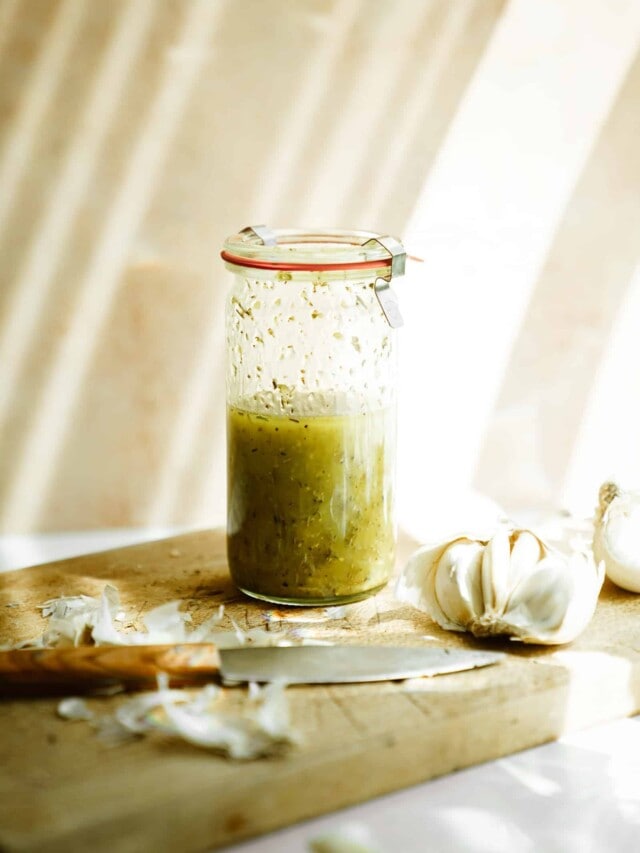 Jar of Greek salad dressing sitting on cutting board with a knife and garlic cloves