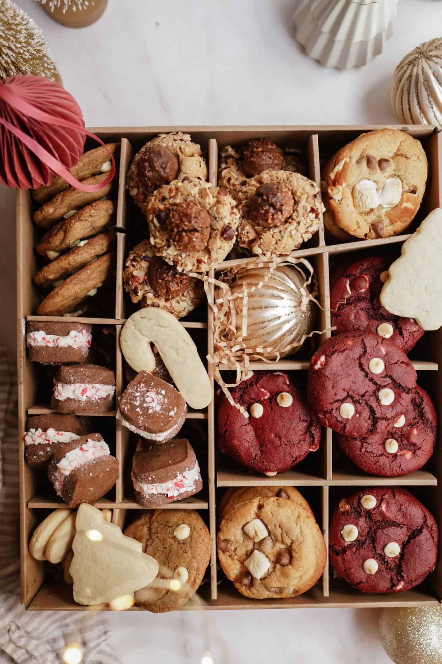 A box filled with a variety of cookies, including red velvet, chocolate chip, marshmallow, and chocolate-covered ones. Some are decorated with nuts or sprinkles. Decor items like paper ornaments and a pinecone are placed around the box.