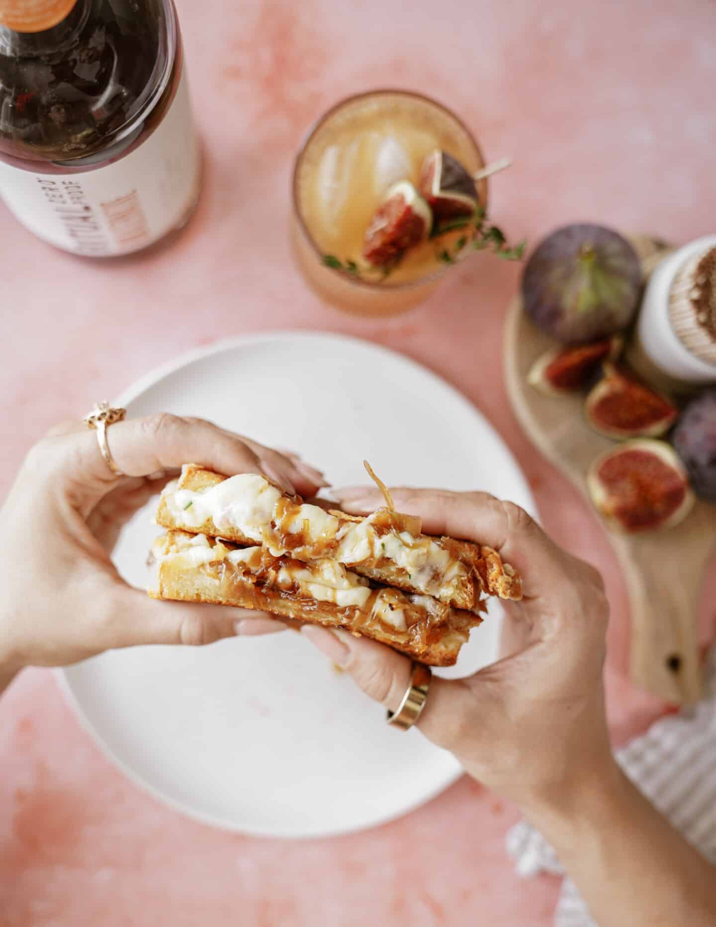 Hands holding a french onion grilled cheese with ingredients around it