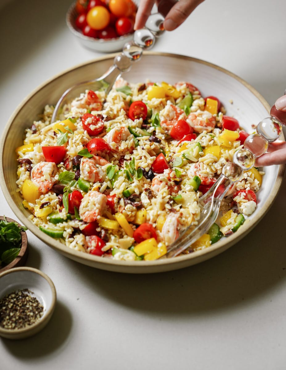 A fresh, colorful salad in a large bowl with shrimp, rice, yellow bell peppers, cherry tomatoes, and green beans. Two hands use clear serving utensils to mix the salad. Small bowls of basil and pepper are nearby on the table.