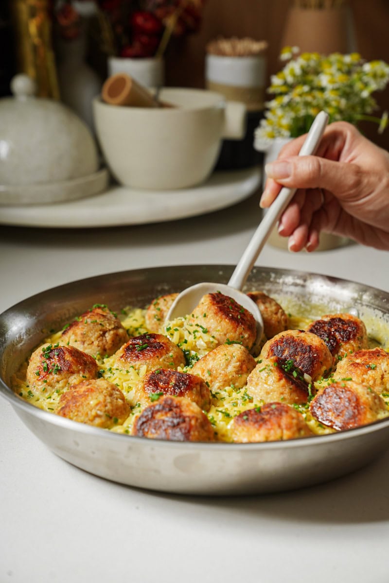 A large grey spoon lifting a golden-brown, perfectly cooked steelhead troutball from a pan. In the background, more fishballs and creamy orzo are visible, showcasing the one-pan nature of the dish.