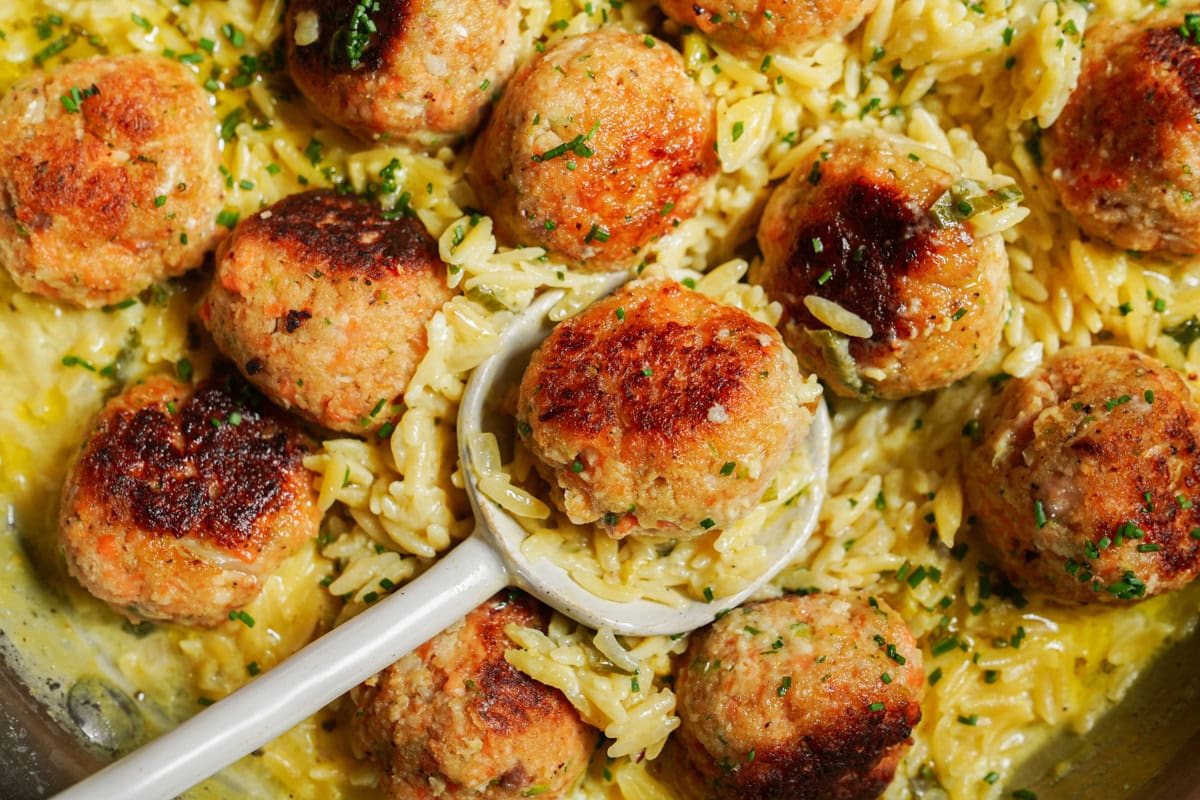 A silver spoon lifts a golden-brown, perfectly cooked steelhead trout ball from a pan filled fish balls and creamy orzo.