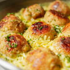 Close-up of sizzling fishballs in a pan. Flecks of herbs and spices are visible on the surface, and the background shows a hint of orzo pasta cooking alongside the meatballs.