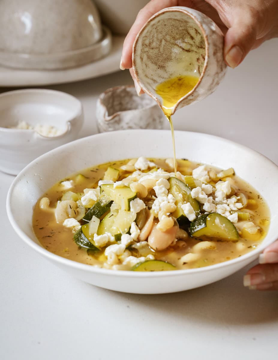 A hand tilts a white cup, pouring broth into a white bowl filled with Butter Bean + Chickpea Noodle Soup. The golden broth cascades over a soup of tender butter beans, chickpea noodles, colorful vegetables, and feta cheese.