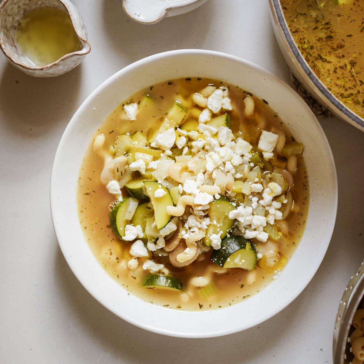 A white bowl filled with Butter Bean + Chickpea Noodle Soup with golden broth, butter beans, twirled chickpea noodles, colorful vegetables, and crumbs of feta.