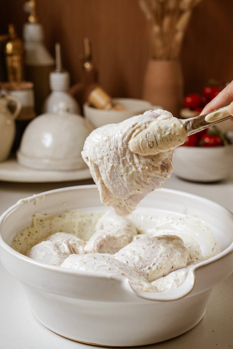 A hand grips a pair of silver tongs, which are clasping a plump chicken thigh coated in a creamy, herb-speckled Greek yogurt marinade. Below it is white ceramic baking dish containing raw chicken pieces coated in the same marinade.  