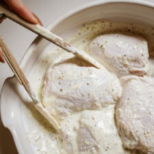 A white ceramic baking dish contains raw chicken pieces coated in a creamy, herb-flecked Greek yogurt marinade.