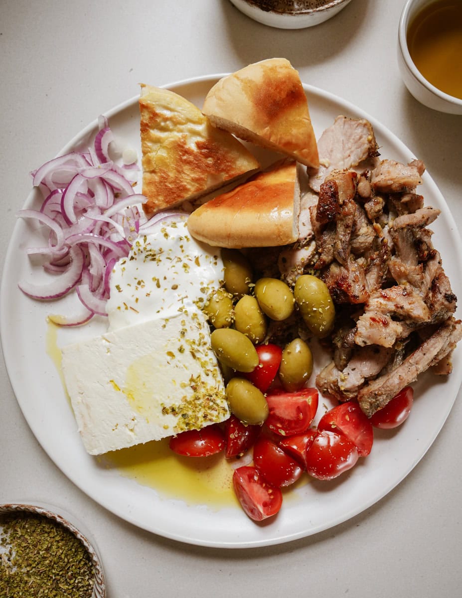 A colorful plate showcases all the elements of a perfect pork gyro: crispy, seasoned pork meat, soft pita bread triangles, briny olives, creamy tzatziki sauce, diced cherry tomoates, a slice of feta cheese, and thin slices of purple onion.