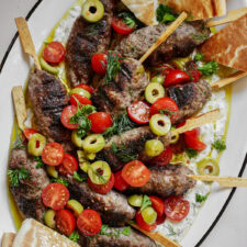 A platter of grilled beef kofta kebabs on tzatziki, topped with tomatoes and olives.