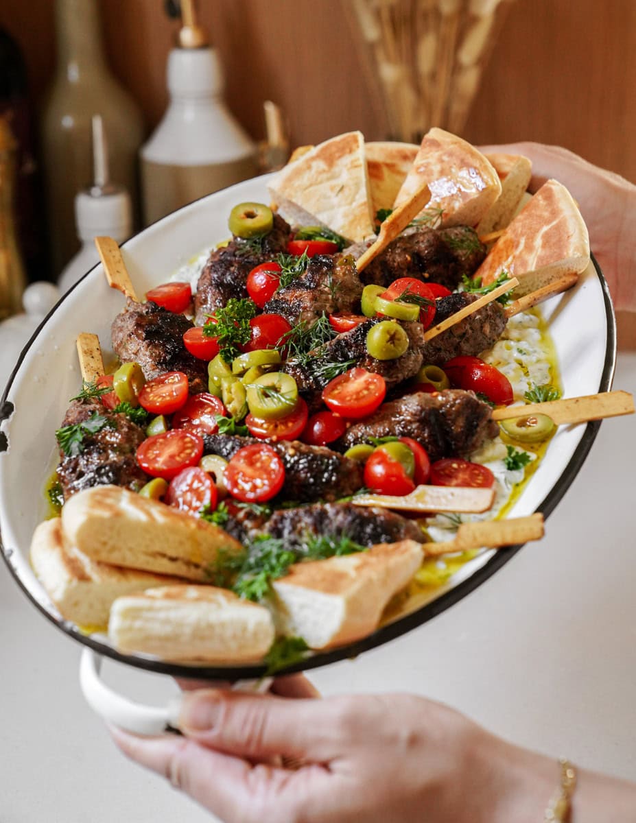 Hands hold up a platter of grilled beef kofta kebabs and pita bread on a bed of tzatziki, topped with tomatoes and olives.