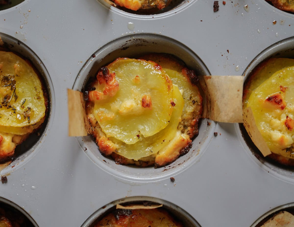 Close-up of baked potato gratin in a muffin tin, featuring thinly sliced potatoes with melted cheese and a golden-brown crust. Each portion is neatly separated in its own cup, with hints of seasoning visible on the surface.