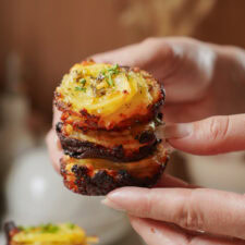 Hands holding a stack of crispy, golden-brown potato slices garnished with herbs and seasoning. The potatoes are thinly sliced and have a roasted texture, appearing appetizing and freshly cooked.