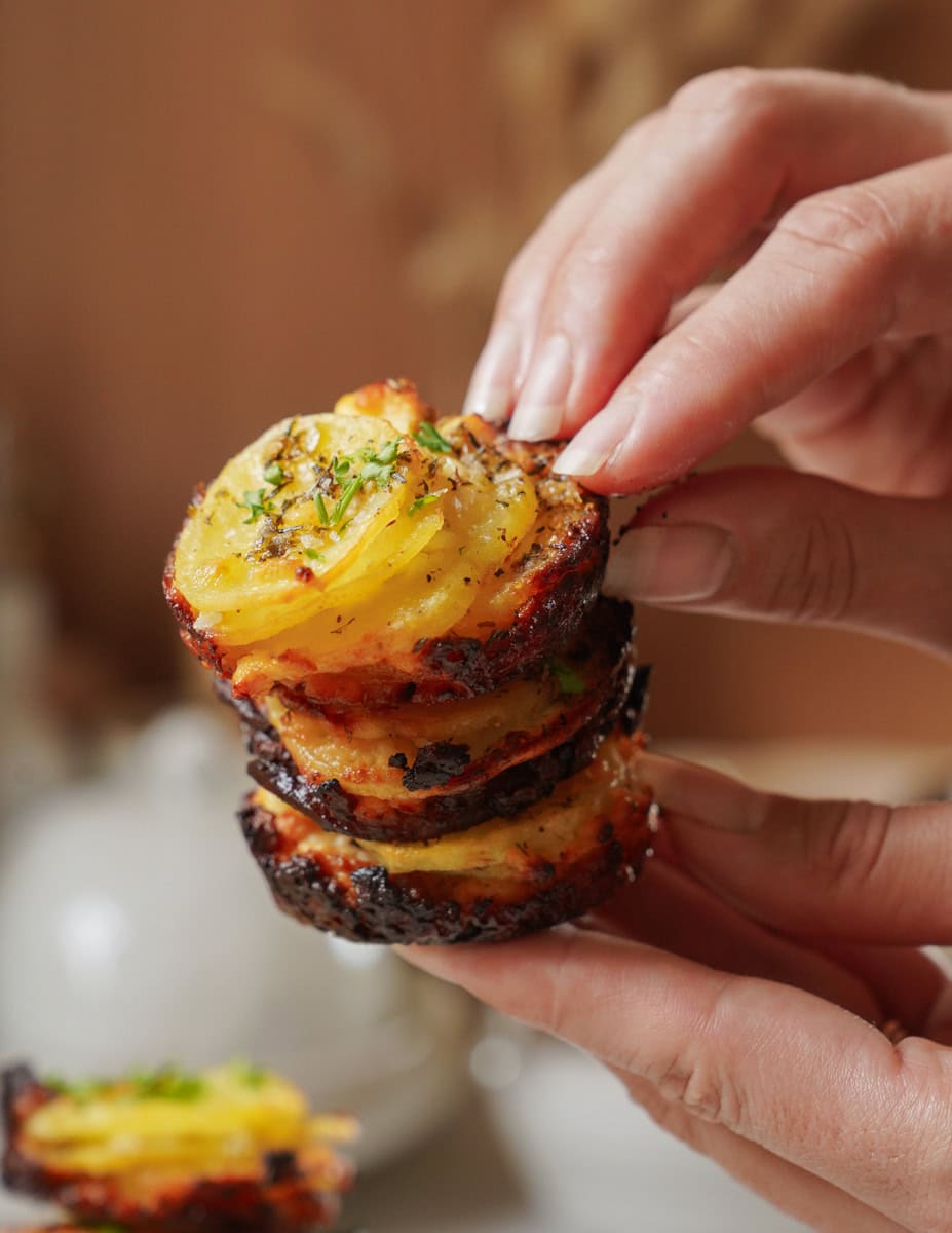 Close-up of hands holding a stack of crispy, baked potato slices garnished with herbs. The potatoes have a golden-brown color, suggesting they are seasoned and flavorful. The background is softly blurred.