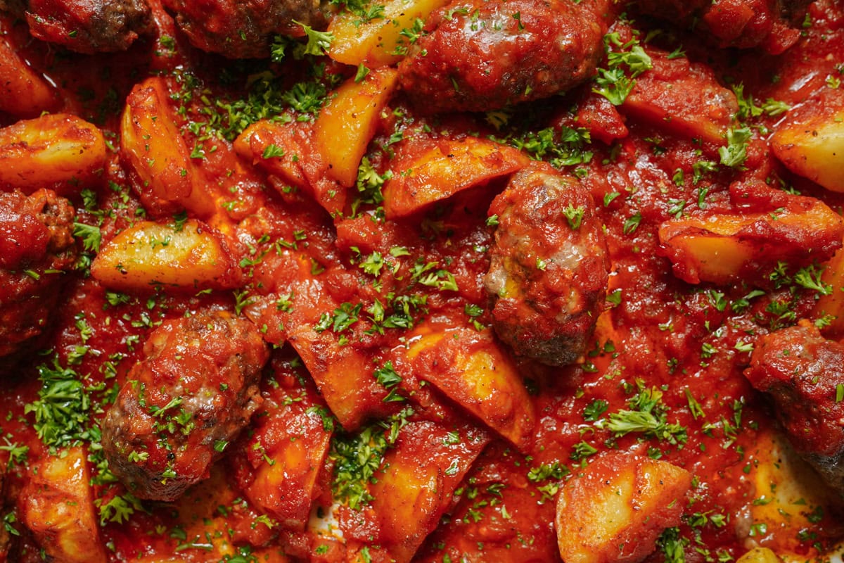 A close-up of a savory dish featuring roasted meatballs and chunks of potatoes coated in rich tomato sauce, garnished with fresh chopped parsley. The vibrant colors highlight the hearty and appetizing texture of the meal.