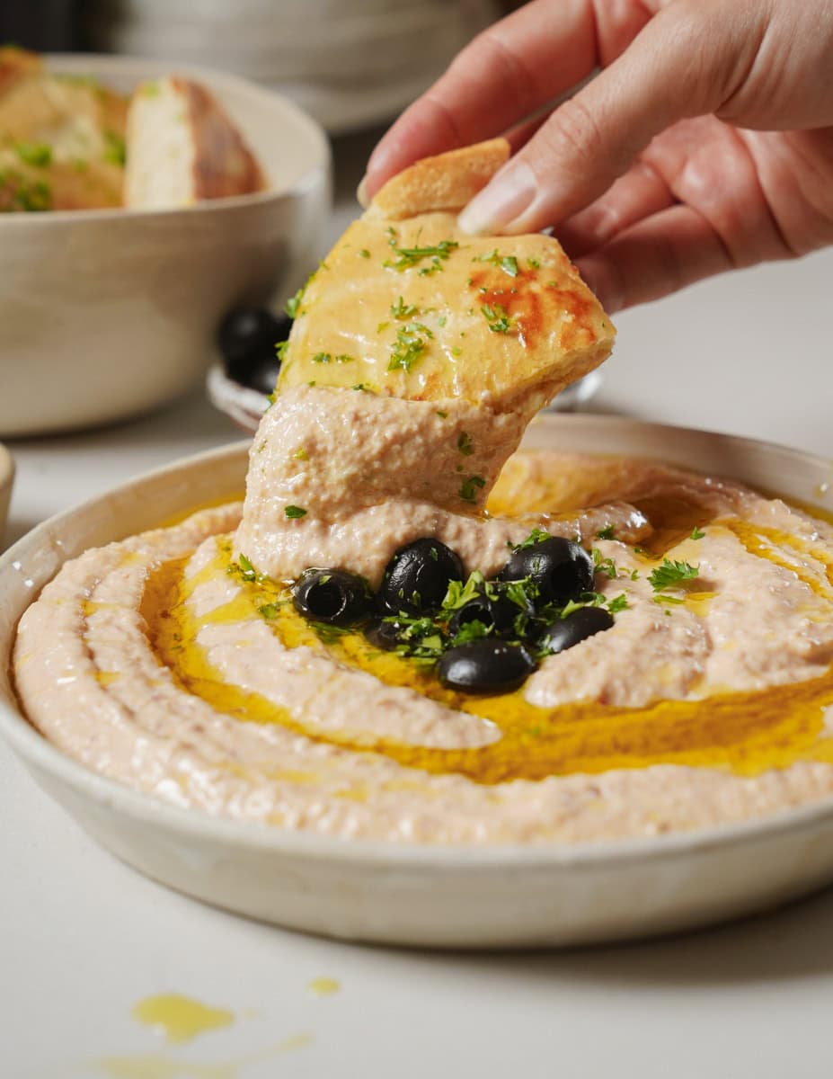 A close-up of a hand dipping a piece of pita bread into a bowl of thick, creamy Taramosalata topped with olives and garnished with fresh herbs. Another bowl with similar contents is partially visible in the background.