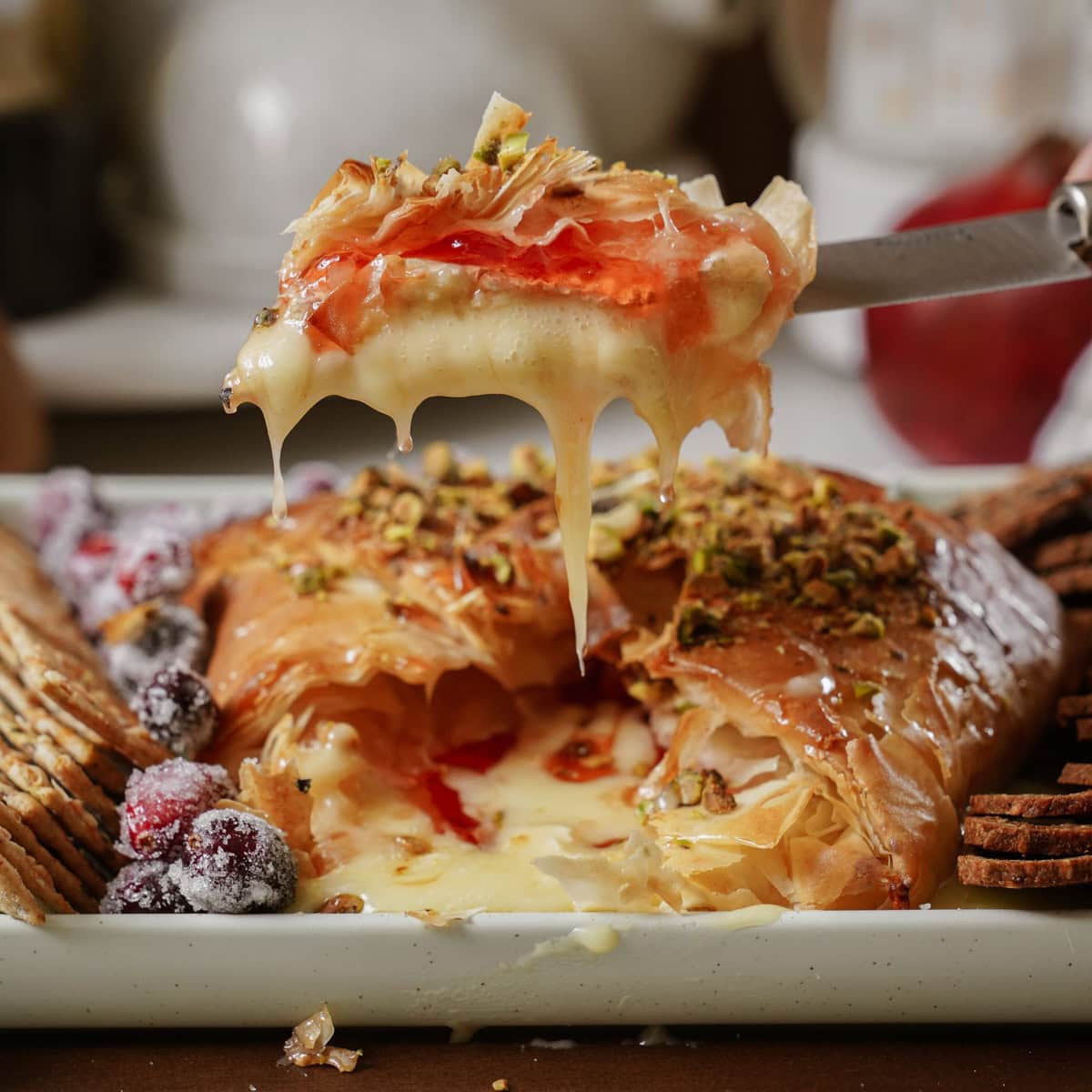 A slice of baked brie wrapped in golden brown phyllo pastry is lifted, showing creamy cheese oozing out and topped with nuts and fruit preserves. Surrounded by crackers and sugared berries on a tray.