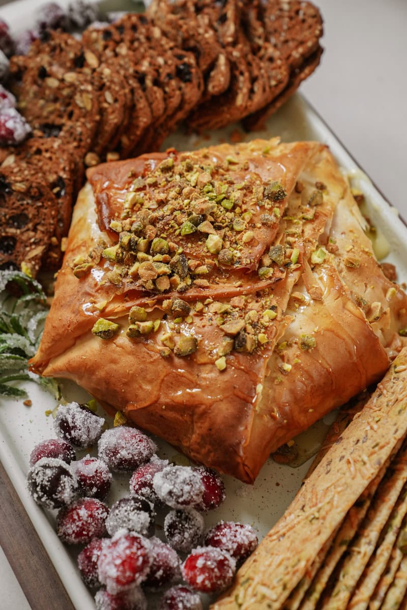 A platter featuring baked brie wrapped in phyllo pastry, topped with chopped pistachios and honey. Surrounded by frosted cranberries, multigrain crackers, and thin breadsticks.