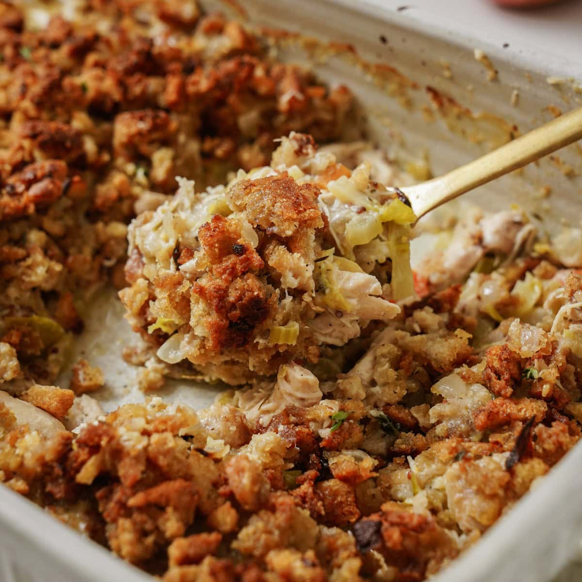 A close-up of a casserole dish filled with a golden-brown stuffing mix. The stuffing is chunky, with pieces of bread, celery, and herbs visible. A spoon is scooping out a portion from the dish.