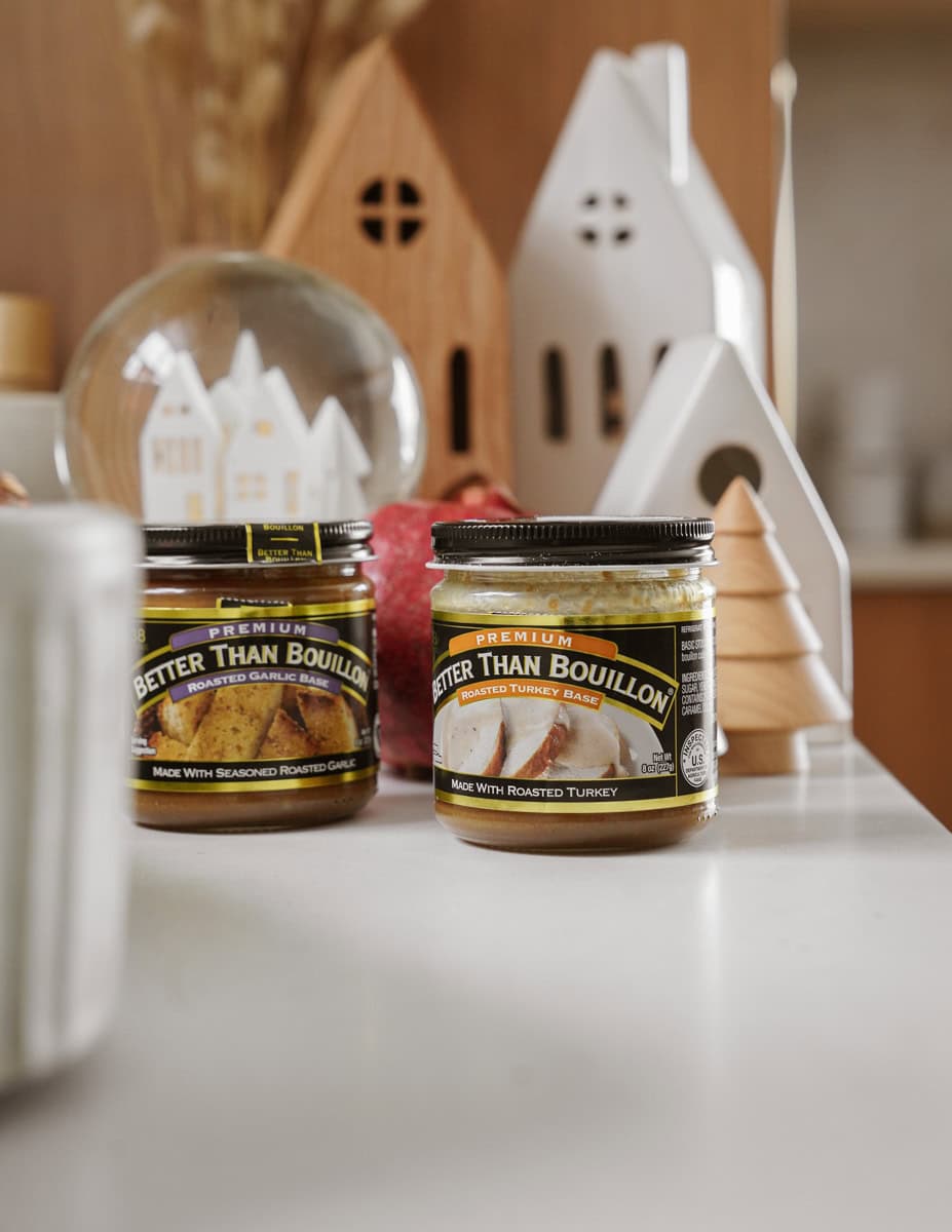 Two jars of Better Than Bouillon sit on a kitchen counter. One is roasted garlic flavor and the other is roasted turkey base. They are surrounded by decorative wooden houses and a small globe with dried plants inside.