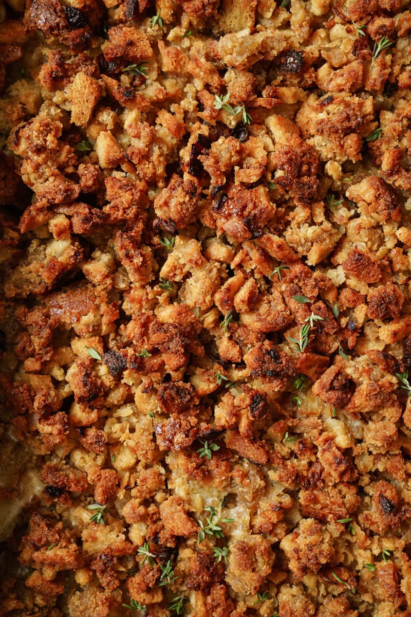 Close-up of golden-brown bread stuffing, with a crispy texture and herbs like thyme sprinkled throughout. The mixture appears moist and baked, showcasing a comforting, homemade appearance suitable for a holiday meal.