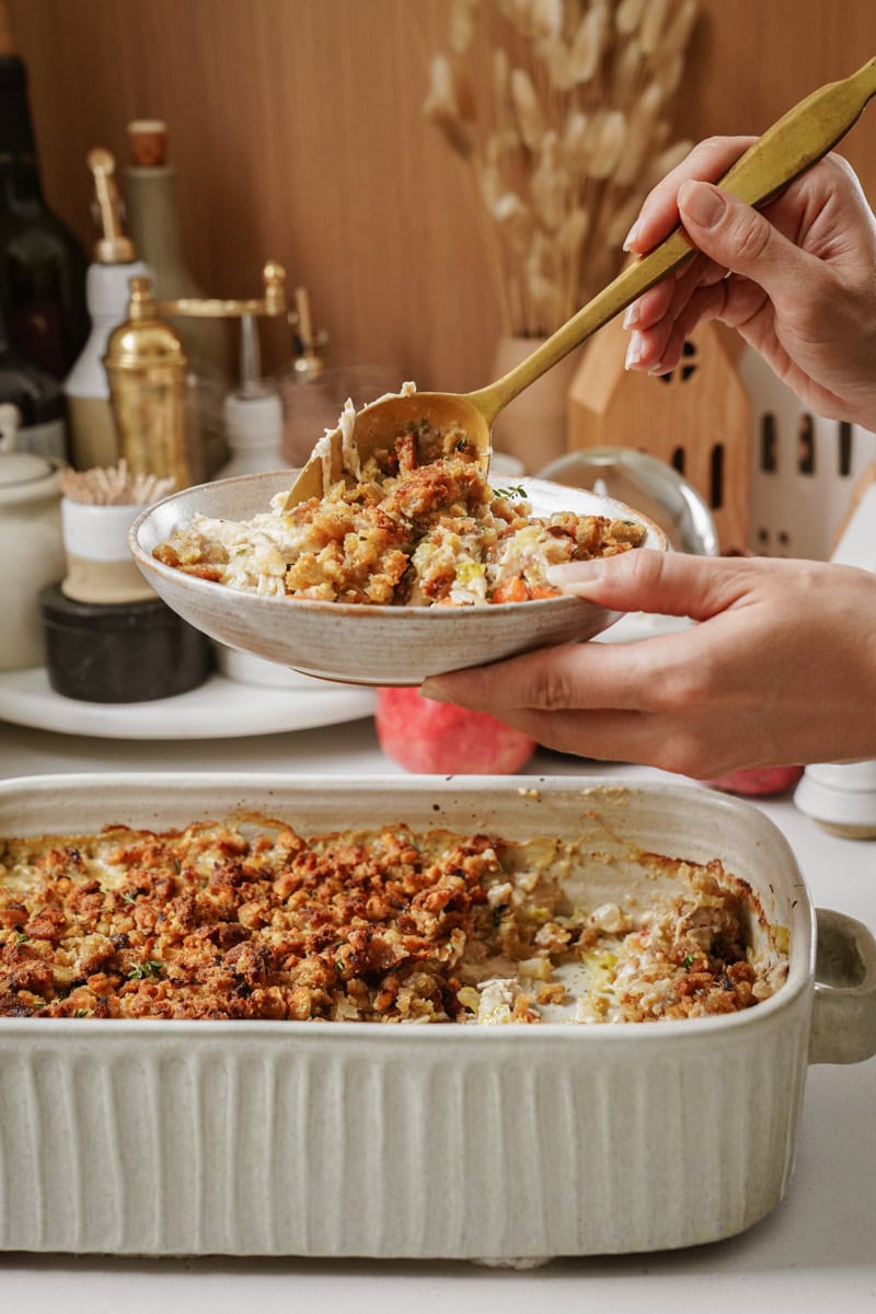 A person is scooping a portion of a baked dish with a crunchy topping from a rectangular ceramic dish into a bowl using a spoon. The kitchen countertop has various utensils and ingredients in the background.