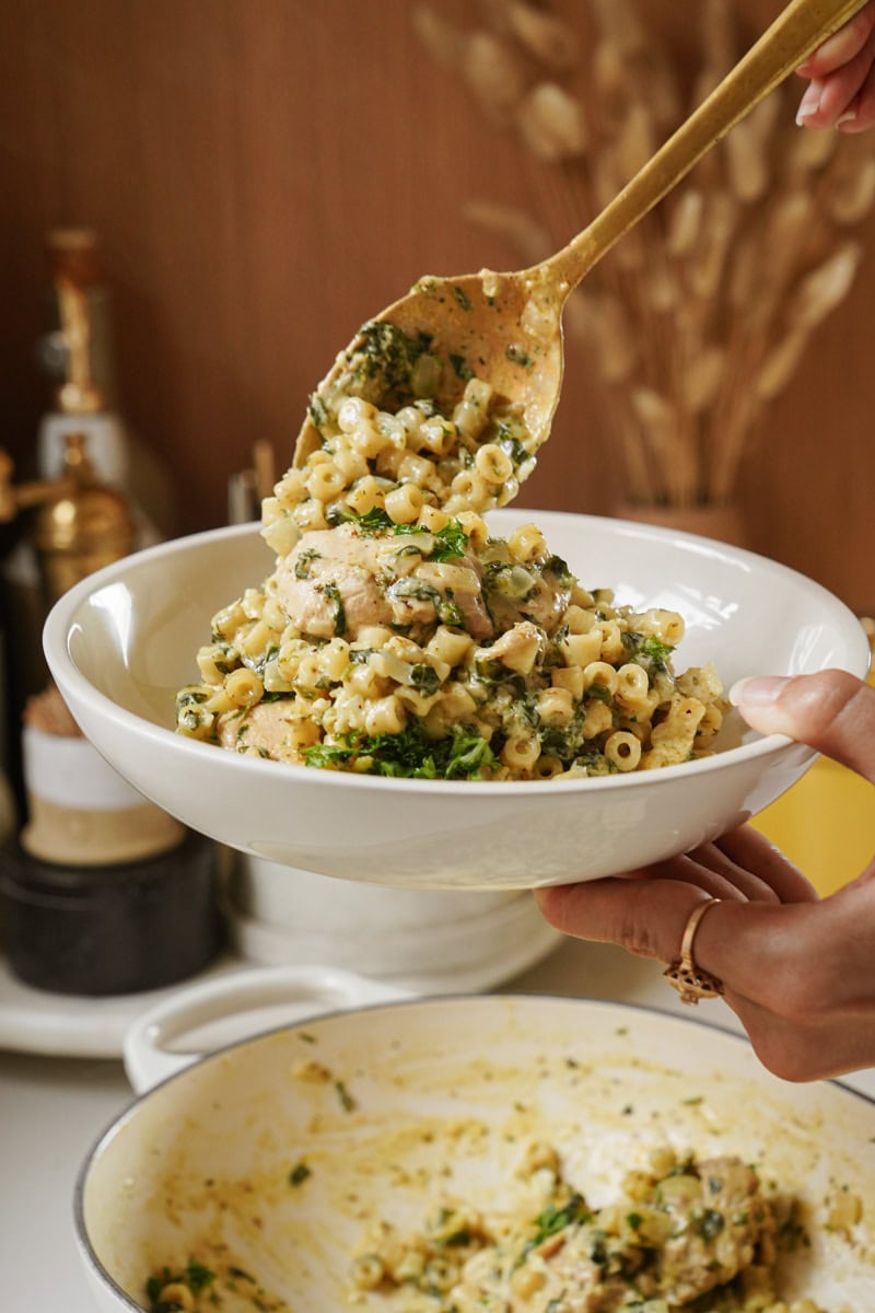 A person serves a dish of pasta in a white bowl using a golden spoon. The pasta is mixed with greens and herbs, creating a colorful presentation. In the background, kitchen containers are visible on a countertop.