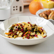 A bowl of creamy yogurt topped with green olives, sliced almonds, pomegranate seeds, and orange zest. In the background, there are small decorative houses, a lit candle, an orange, and a plate of sliced bread.