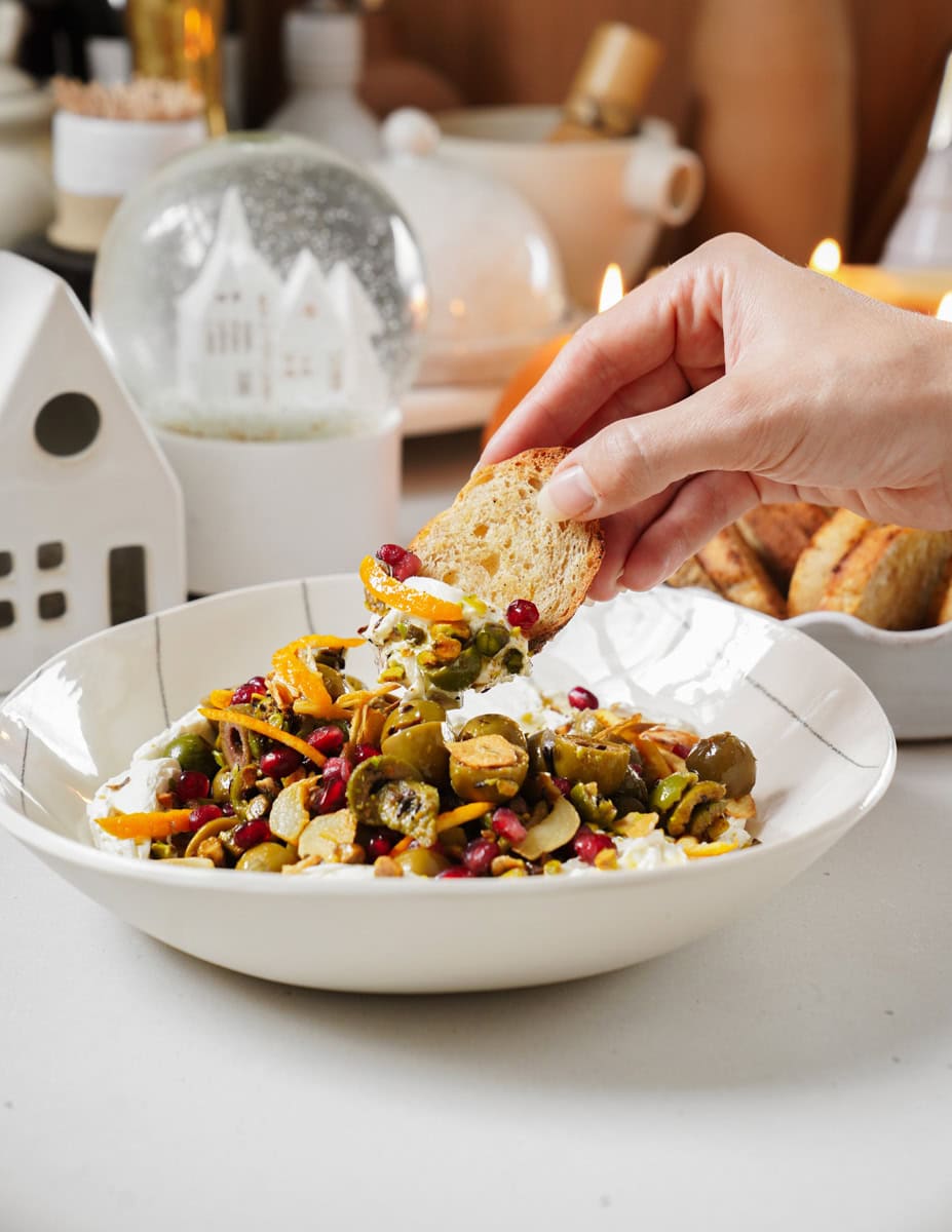 A hand holds a slice of bread dipped into a bowl filled with a colorful mixture of green olives, pistachios, herbs, and pomegranate seeds. The background features cozy, decorative items like a snow globe and candles.