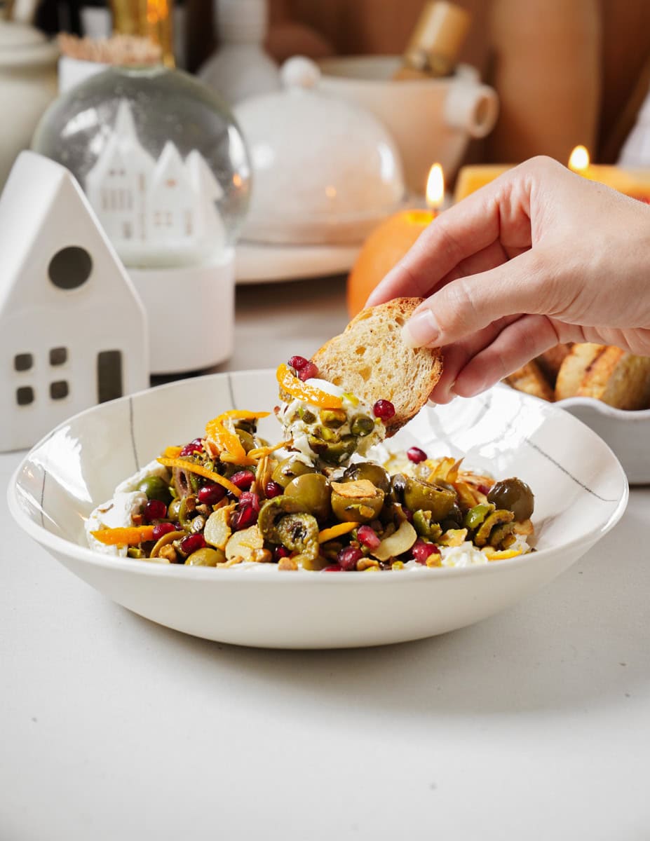 A hand is dipping a piece of bread into a salad bowl filled with mixed vegetables, pomegranate seeds, and olives. The table has a cozy, decorative setting with a lit candle, a snow globe, and a small house ornament in the background.