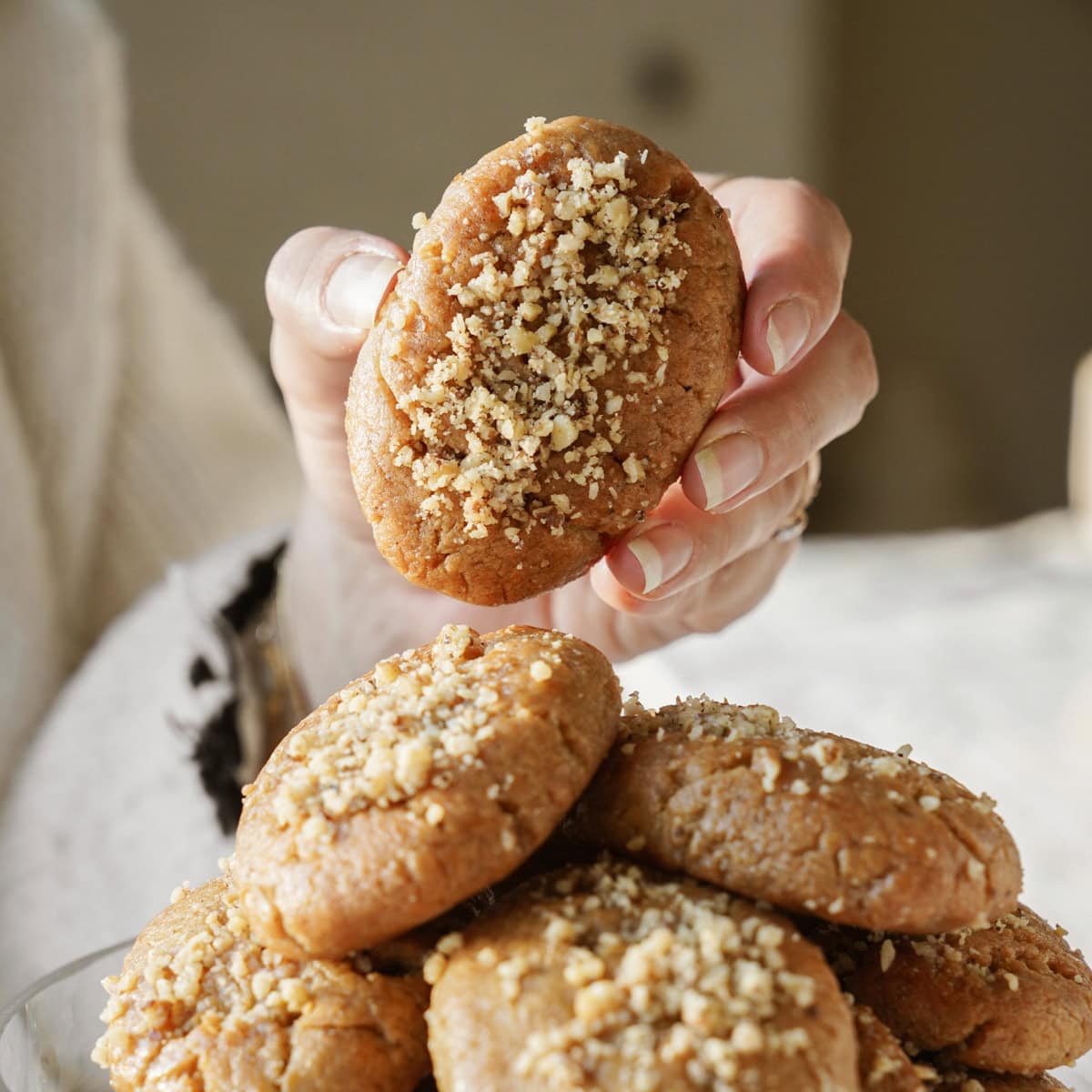 A person holding a cookie topped with crushed nuts, with more cookies in a pile below. The background is softly blurred, focusing attention on the homemade treats.