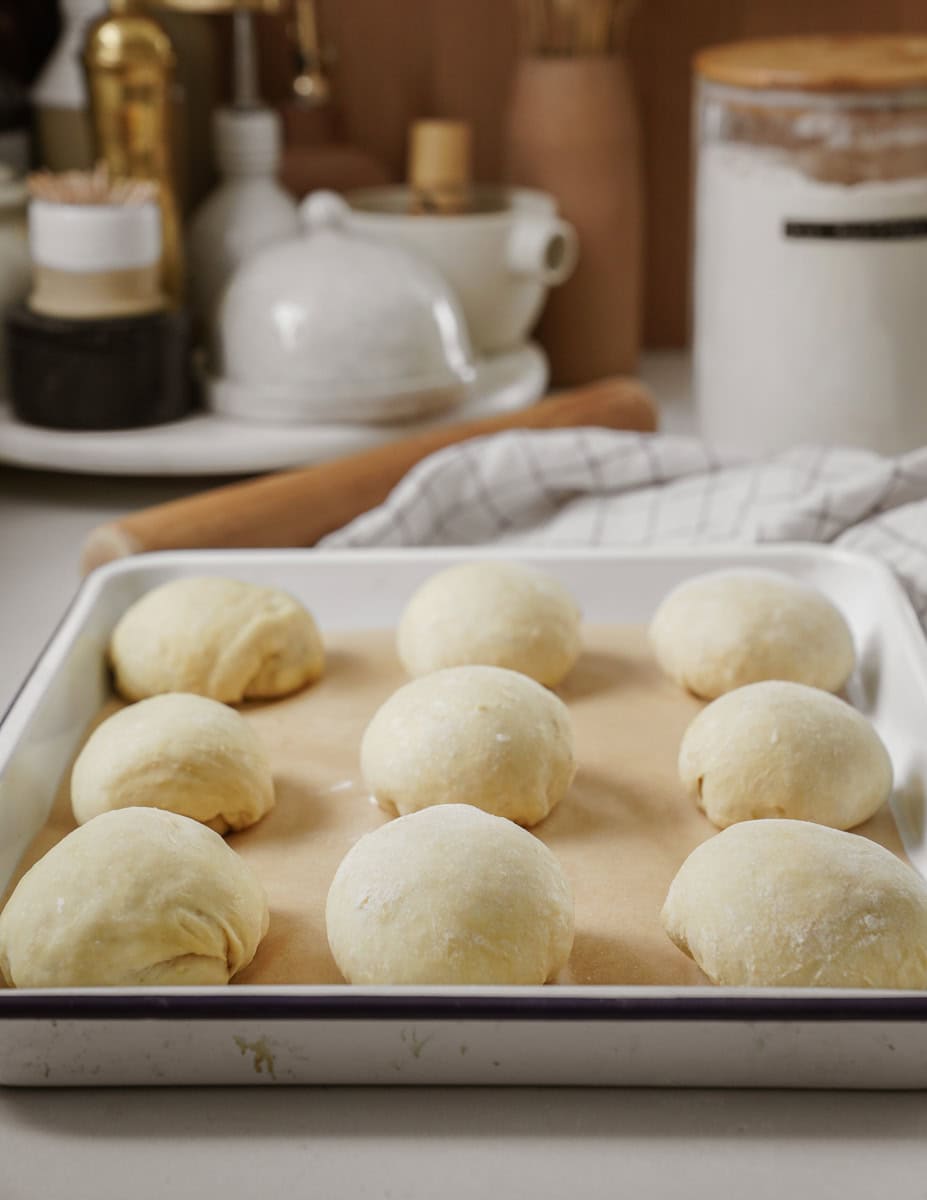 Nine round dough balls are neatly arranged on a parchment-lined baking tray. In the background, kitchen items like a rolling pin, a jar of flour, and a teapot are visible, adding a homely culinary atmosphere to the scene.