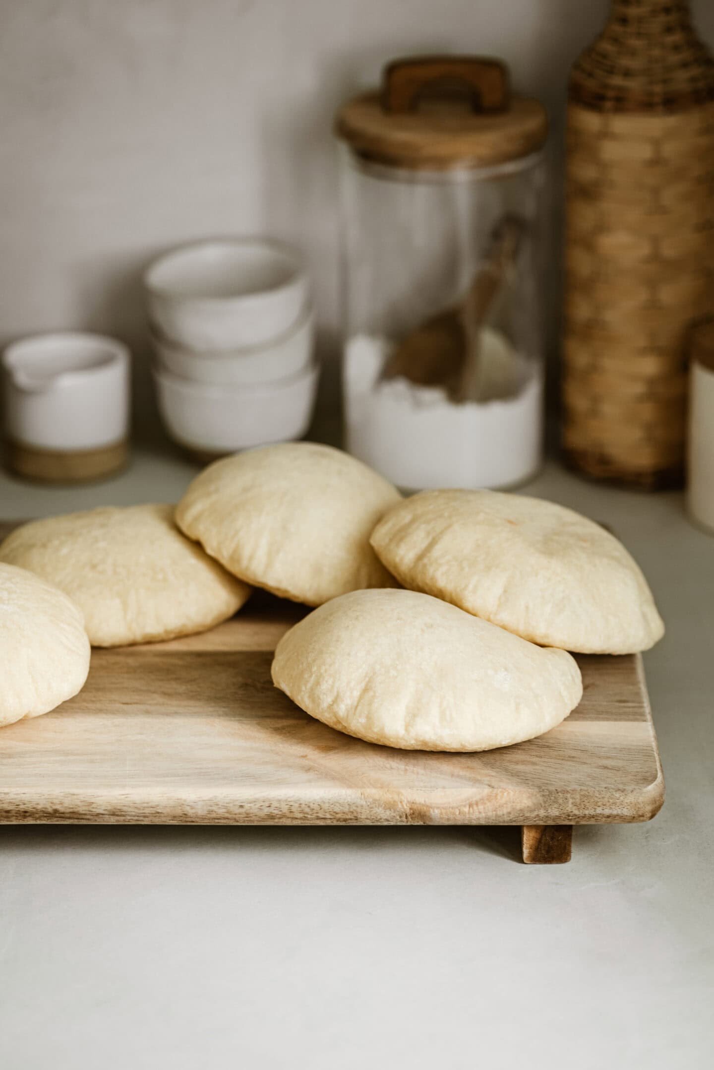 A wooden tray holds several freshly baked pita breads in a cozy kitchen setting. In the background, there are white bowls, a jar of sugar, and a wicker container, adding to the rustic ambiance.