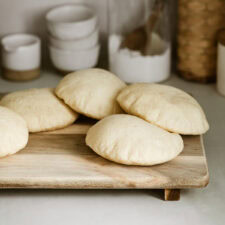 Five freshly baked pita breads are placed on a wooden board in a kitchen setting. In the background, there are white bowls, a small pitcher, a jar with a wooden spoon, and a wicker basket, all on a light-colored countertop.