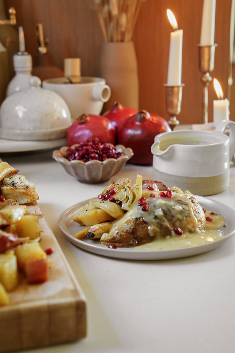 A cozy table setting with a plate of roasted chicken topped with pomegranate seeds and creamy sauce. Nearby, sliced fruit, whole pomegranates, candles, and pottery add warmth to the scene.