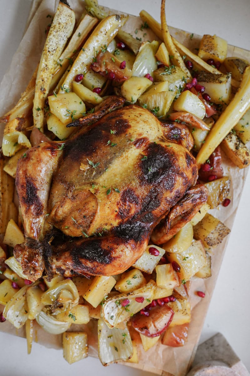 A roasted chicken with a golden skin is placed on parchment paper surrounded by roasted vegetables. Vegetables include potatoes, onions, and carrots, garnished with herbs and pomegranate seeds.