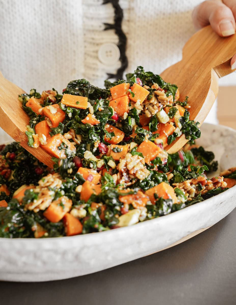 A person tosses a colorful salad with wooden salad tongs in a white bowl. The salad includes kale, yams, seeds, nuts, and pomegranate seeds. The person is wearing a white sweater with a visible button.