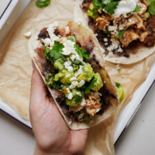 A hand holding a taco filled with chicken, black beans, avocado, cheese, cilantro, and sour cream. Another taco can be seen on a tray in the background, resting on parchment paper.