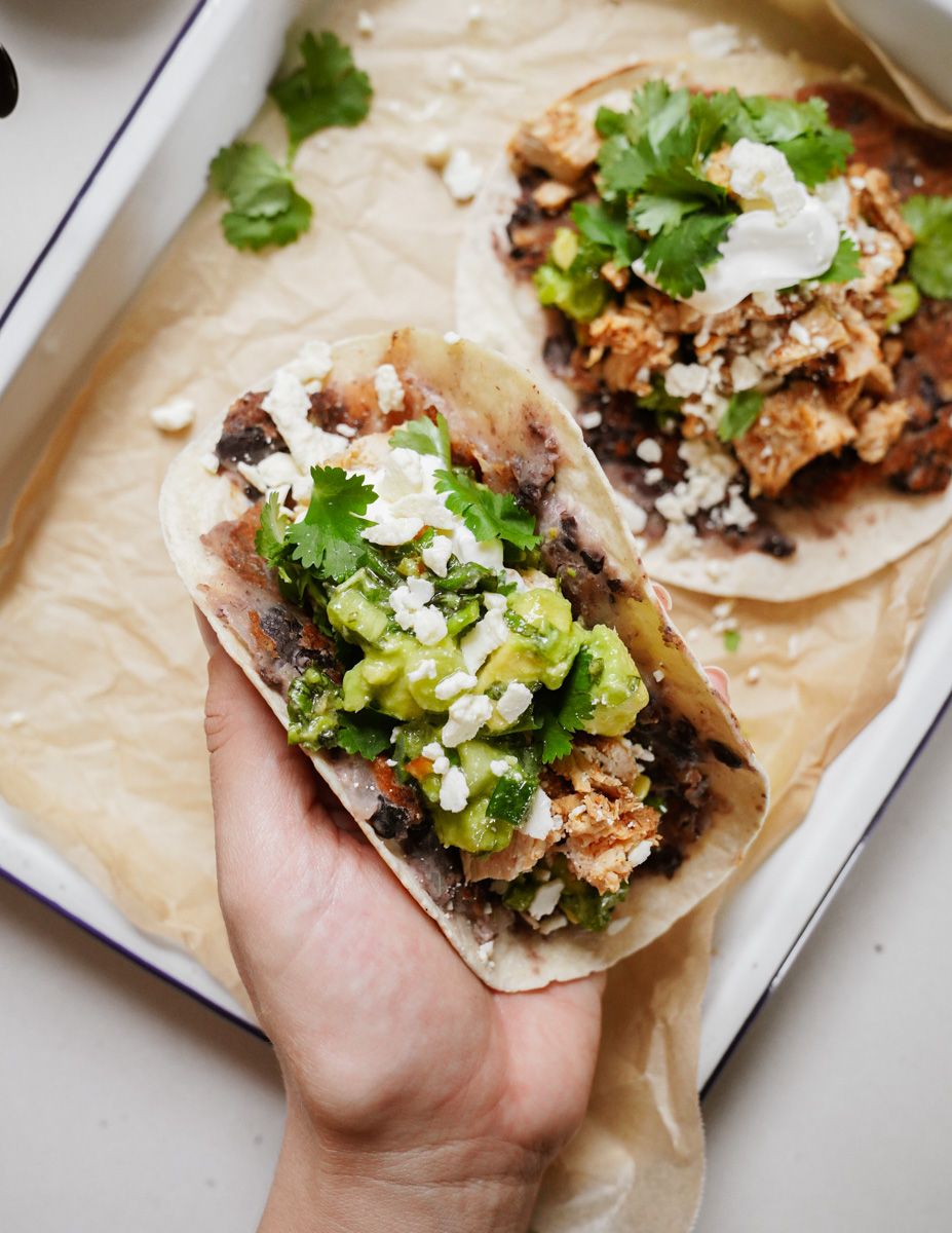 A hand holds a taco filled with black beans, chicken, avocado, cilantro, and crumbled cheese. Another taco with similar ingredients is visible on a white paper-lined tray in the background.