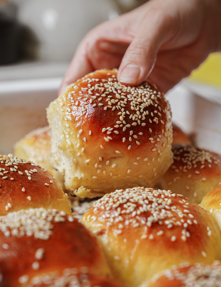 A hand picking up a freshly baked, golden-brown sesame seed bun from a tray. The buns are glossy and arranged closely together, with a soft, fluffy texture visible in the torn piece.