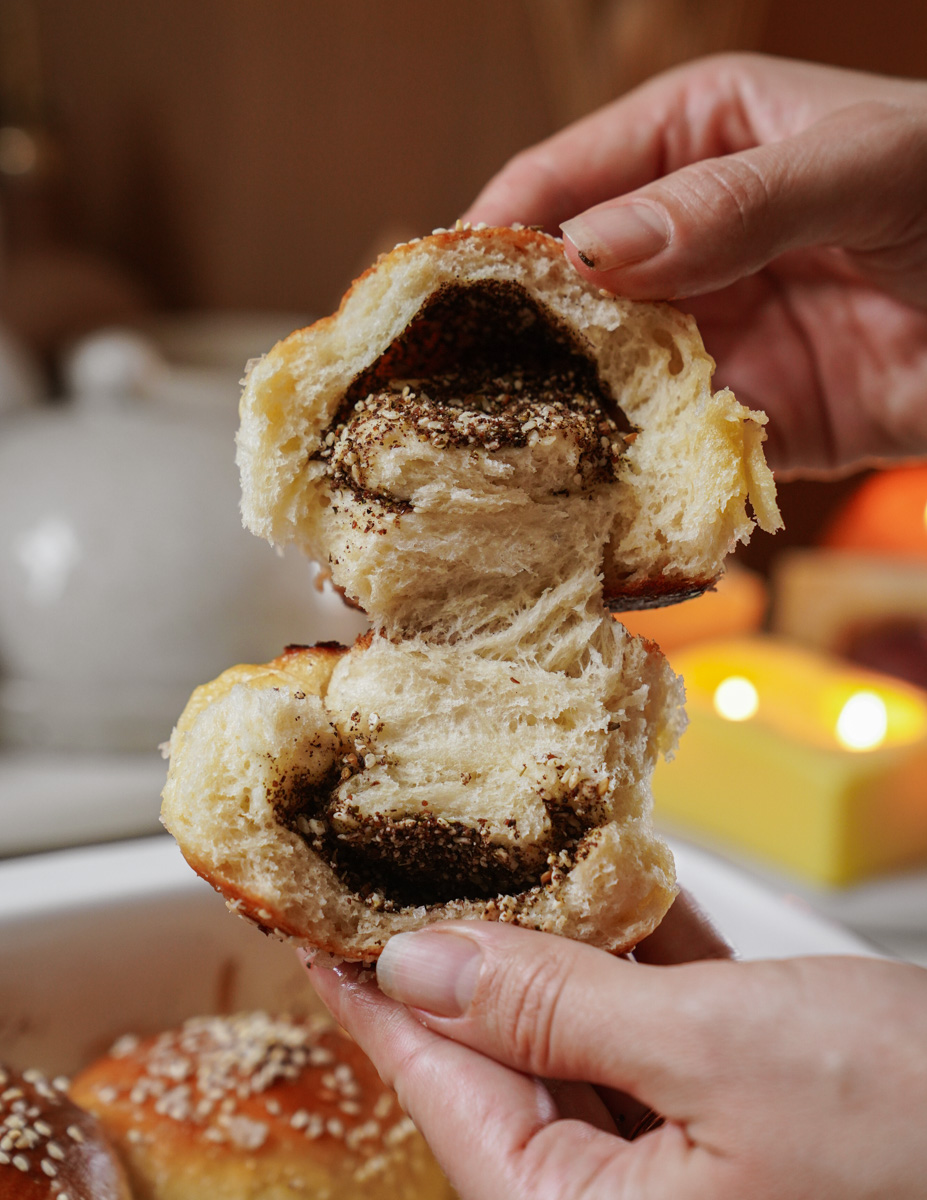 Hands pulling apart a soft, sesame-topped bread roll filled with chocolate spread, with candles and kitchenware blurred in the background.