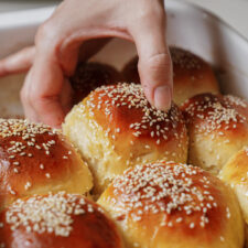 A hand reaches into a baking dish to pick up a freshly baked, golden brown dinner roll topped with sesame seeds. The rolls are closely packed together, each with a shiny, glazed finish.