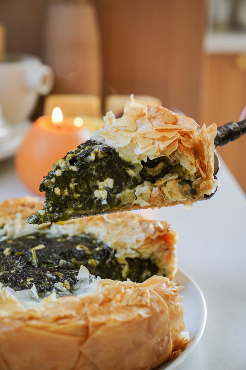 A slice of Greek spinach pie being lifted from a whole pie on a plate. The pie has a flaky, golden-brown crust and a rich green filling, likely spinach and cheese. Candles and soft lighting create a cozy atmosphere in the background.