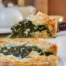 A slice of spinach pie is being served from a whole pie. The pie has a flaky, golden crust and is filled with a dark green spinach mixture containing cheese. The background is softly blurred, showing a white teapot.