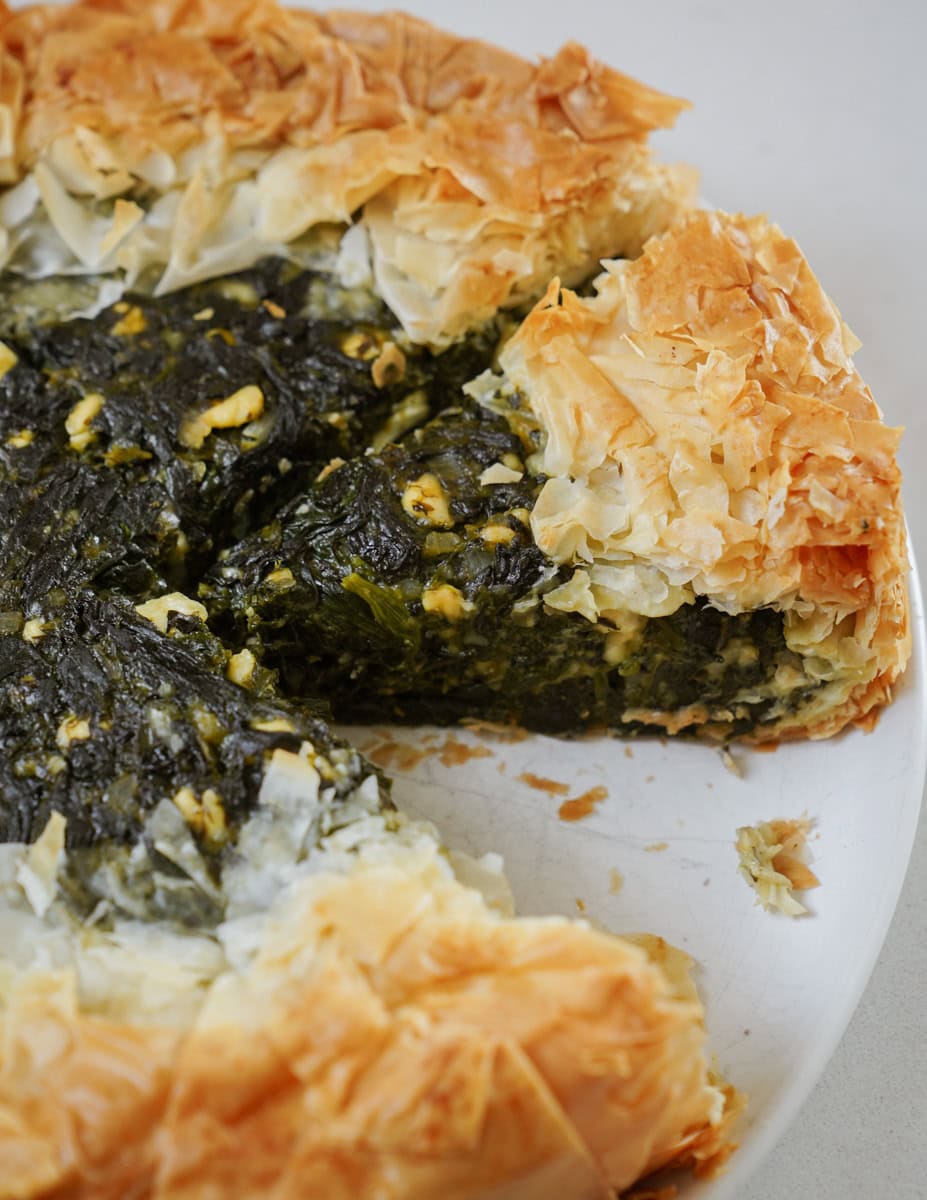 A close-up of a spinach and cheese pie on a white plate. The pie has flaky, golden-brown crust edges, with a dark green spinach filling visible in the removed slice section.